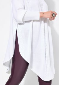 Witte gebreide poncho met een high-low zoom, voorzien van textuurrijke kabelbrei-details langs de zijkanten. Gepaard met nauwsluitende bordeauxrode leggings.