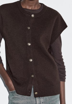 Gilet sans manches marron foncé boutonné porté sur un haut à manches longues, associé à un jean bleu clair, main dans la poche.
