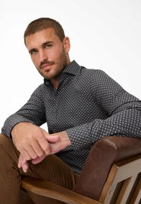 Dunkel gemustertes Hemd mit geometrischen Mustern in Creme und Blau, aus atmungsaktivem Stoff, sitzt auf einem Holzstuhl.