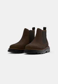 Botoșei Chelsea din suede maro, cu panouri elastice laterale, vârf rotund și sole din cauciuc negru. Dispune de tab-uri pentru o purtare ușoară.