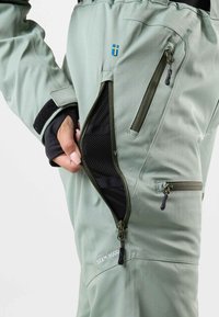 Green water-resistant pants with zippered pockets, mesh lining, and adjustable waistband. Features logo, contrasting zipper details, and sturdy fabric.