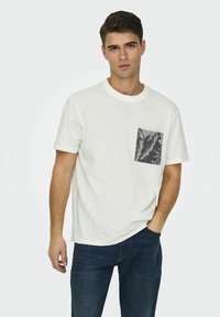 T-shirt bianco in cotone con scollo rotondo, caratterizzato da una tasca a motivo foglia in bianco e nero sul lato sinistro. Abbinato a jeans scuri.