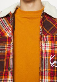 Koszula flanelowa w kratę z kołnierzem sherpa w odcieniach czerwieni, pomarańczy i brązu, noszona na żółtej koszulce z krótkim rękawem i okrągłym dekoltem.