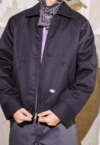 Svart zip-up jacka med spetsig krage, sidfickor och en slät yta. Har en framträdande varumärkesetikett på den nedre framsidan.