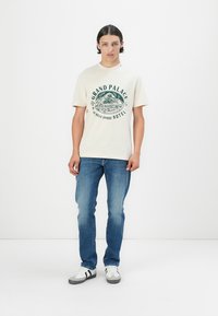 Tricou bej din bumbac cu imprimeu verde care spune "GRAND PALACE, 17 Belle Epoque HOTEL." Purtați cu blugi albaștri și adidași albi cu dungi negre.