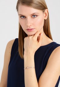 Bracelet en argent avec un accent en or rose circulaire, présentant un design tissé. Le modèle porte un haut bleu foncé, la main posée sur le menton.