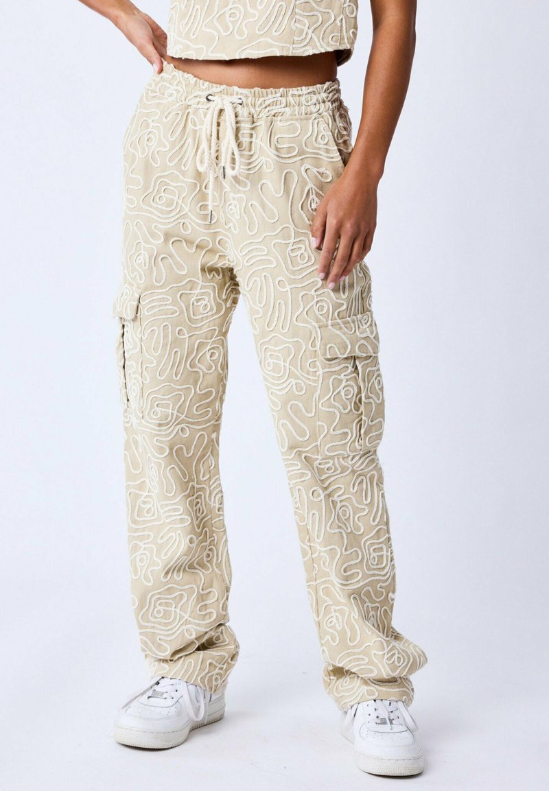Pantaloni cargo beige con motivi astratti bianchi, vita elasticizzata con coulisse, tasche laterali e una vestibilità rilassata. Indossati con sneakers bianche.