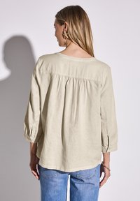 Camicia di lino beige chiaro con yoke posteriore e pieghe, maniche a tre quarti con polsini a bottone, abbinata a jeans in denim blu.