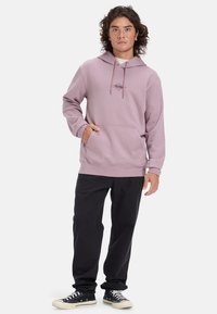 Sudadera ligera de color púrpura con un bolsillo tipo canguro, capucha con cordón y logo bordado. Combinada con pantalones negros y zapatillas negras.