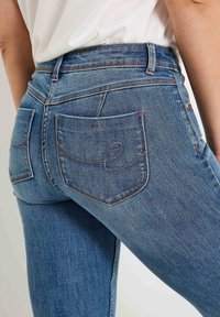 Jean en denim bleu foncé avec une coupe ajustée, présentant deux poches arrière avec des détails de couture courbés et une taille avec passants de ceinture.