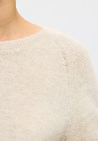 Primer plano de una persona que lleva un suéter de punto suave de color crema con cuello redondo sobre un fondo liso y claro.