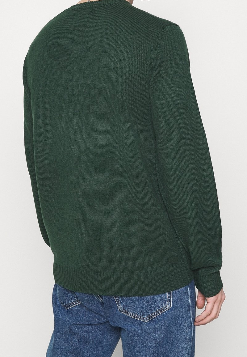 Homme portant un pull en maille vert foncé et un jean en denim bleu, vu de dos sur un fond clair uni.