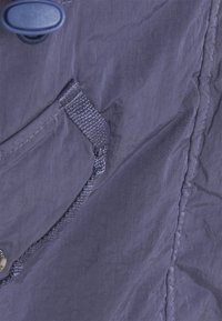 Lila lättviktsjacka med strukturerad tyg, framficka med webbing och metallknappsstängning på höger sida.