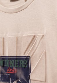 Beige katoenen t-shirt met tekst met reliëfletters. Voorzien van een grafische print met "WINNAARS" in groene en rode accenten op een blauwe achtergrond.