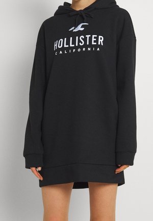 Osoba nosząca za dużą czarną bluzę z kapturem Hollister California z białym logo i kapturem, zakrywającą górne partie nóg na jednolitym tle.