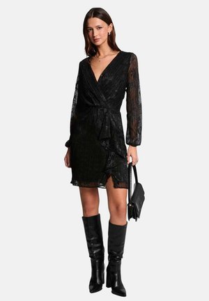 Vestido negro de manga larga, tipo wrap, con escote en V, dobladillo con volantes y textura transparente, combinado con botas negras hasta la rodilla y un bolso pequeño.