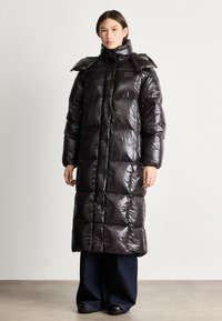 Caban lung din puf negru, cu un finisaj lucios, matlasat, guler înalt și un design de umăr supradimensionat. Dispune de închidere cu fermoar și buzunare laterale.