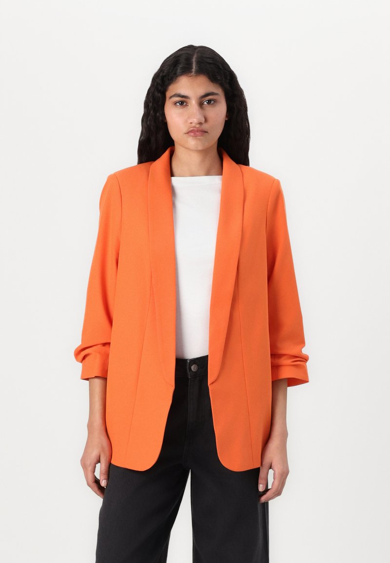 Orange blazer med skræddersyet pasform, notched klapper og trekvartærmer, parret med en hvid top og sorte bredbenede bukser.