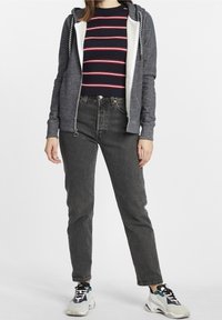 Sudadera gris oscuro con cremallera y cordón blanco, superpuesta sobre una camiseta a rayas azul marino. Jeans gris claro y zapatillas multicolores completan el look.