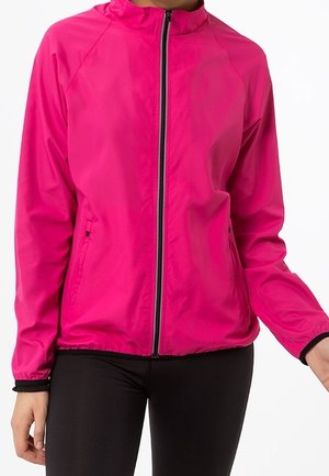 Žena v jasně růžové bundě na zip s stojáčkem a černým zipem, v kombinaci s černými legínami, zobrazena od krku po boky.