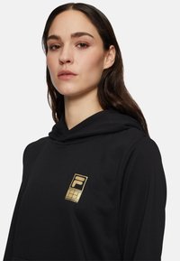 Svart hoodie med en guldig Fila-logotyp på bröstet. Mjuk material med dragsko vid huvan och en slät, minimalistisk design.