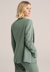 Zelený přizpůsobený blazer s hladkým povrchem, jednoramenným designem a klopami. Obsahuje strukturovaný střih a boční kapsy.