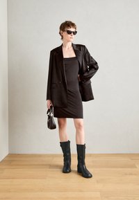 Černý přizpůsobený blazer přes upnuté černé šaty, doplněný černými holínkami po kolena. Doplňky zahrnují malou kabelku a sluneční brýle.