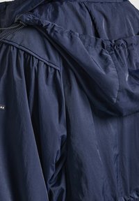 Námořnický modrý kabát s kapucí vyrobený z lehkého materiálu s nařasením na ramenou a manžetách. Obsahuje dekorativní nášivku s logem.