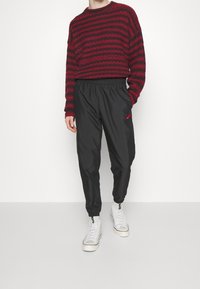 Red and black striped knitted sweater, paired with black tapered joggers featuring an elastic waistband and small red logo, worn with white sneakers.