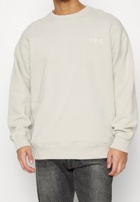 Svetle béžová mikina s kulatým výstřihem a spadlými rameny. Na hrudi se nachází logo YPS v jemném vyšívání. Měkká textura, ležérní střih.