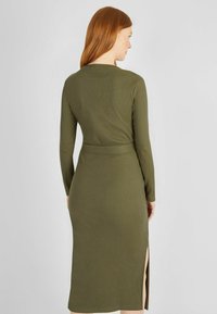 Olivgrünes, geripptes Kleid mit langen Ärmeln, figurbetonter Silhouette, tailliertem Gürtel und seitlichem Schlitz für zusätzliche Bewegungsfreiheit.