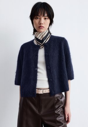 Navy blue short-sleeve cardigan made of fuzzy material with round buttons. Worn over a white top and paired with brown leather pants.