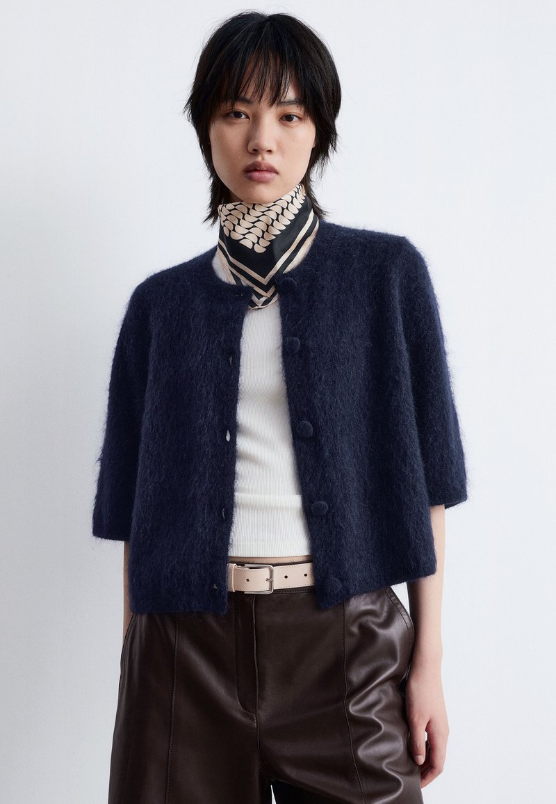 Cardigan bleu marine à manches courtes en matière douce avec des boutons ronds. Porté sur un haut blanc et associé à un pantalon en cuir marron.