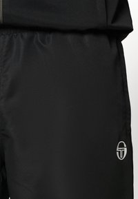 Fekete sportnadrág texturált anyagból. Rugalmas derékpánttal és ezüstszínű logóval a jobb alsó részen.