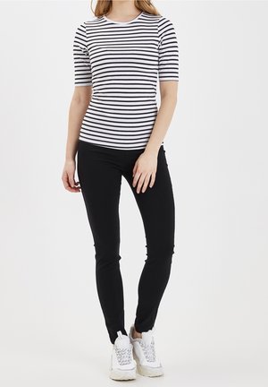 Kobieta ubrana w dopasowaną czarno-białą koszulkę w paski na krótki rękaw, czarne obcisłe spodnie oraz białe masywne trampki, stojąca z jedną ręką swobodnie opuszczoną.