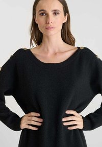 Maglione nero lavorato a maglia con scollatura ampia, maniche lunghe e dettagli in bottoni dorati sulle spalle. Texture morbida e vestibilità rilassata.