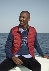 Gilet trapuntato rosso sopra una felpa blu con logo bianco, sovrapposto a una camicia a righe blu e bianche, abbinato a pantaloni di velluto corduroy scuri.
