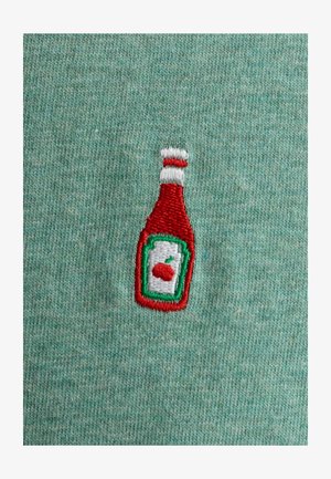 Geborduurd ketchupflesmotief in rood, wit en groen op een gestructureerde mintgroene stofachtergrond.