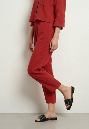 Person iført teksturerede røde bukser og matchende top, stående på ét ben, med sorte slidesandaler med store sølvspænder.