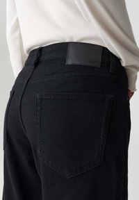 Jeans in denim nero con una texture liscia e design a cinque tasche. Presentano una patch in pelle sulla parte posteriore della vita e cuciture a contrasto.