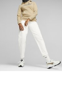 Person som bär en beige huvtröja, vita byxor, vita Puma-strumpor och vita sneakers med leopardmönster som står mot en grå bakgrund.