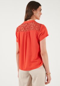 Blusa rossa a maniche corte con dettagli intricati in crochet sul collo posteriore, morbido al tatto e vestibilità rilassata con orlo arrotondato.