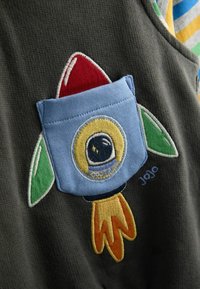 Pilkas audinys su įsiūtu raketos dizainu, raudonu galiuku, mėlyna kišene, žaliomis uodegomis ir geltonais akcentais, kuriuose gausiai išsiuvinėta siuvinėjimas.