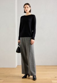 Zwarte gebreide trui, grijze wijde broek met een textuurpatroon, en zwarte hakken. Het model houdt een kleine zwarte handtas vast.