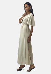 Long, cream-colored dress with short puff sleeves, smocked waist, and flowing fabric. Paired with black strappy heels and silver accessories.