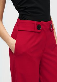 Naine, kes kannab kõrge vöökohaga punaseid pükse mustade nuppudega ja musta kogulist topi, käsi püksitaskus.