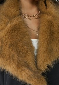 Bruine faux bontkraag op een donkere jas, met gelaagde gouden kettinkjes met een kruisvormige hanger en een wit topje eronder.