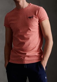 Röd texturerad bomulls-t-shirt med korta ärmar och rundad halsringning, med en liten blå logo på bröstet, kombinerad med mörka byxor.