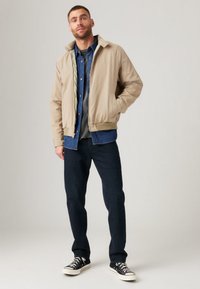 Beige bomber jacket with a collar, zip closure, and elastic cuffs, layered over a denim shirt, paired with dark jeans and black sneakers.