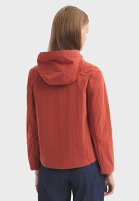 Veste à capuche rouge en tissu texturé, avec une coupe droite et des manches longues. L'ourlet est légèrement arrondi et se termine à la taille.